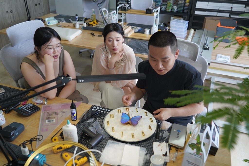 三位工坊伙伴围桌协作，细致组装以蓝色闪蝶为中心的贝壳钟面