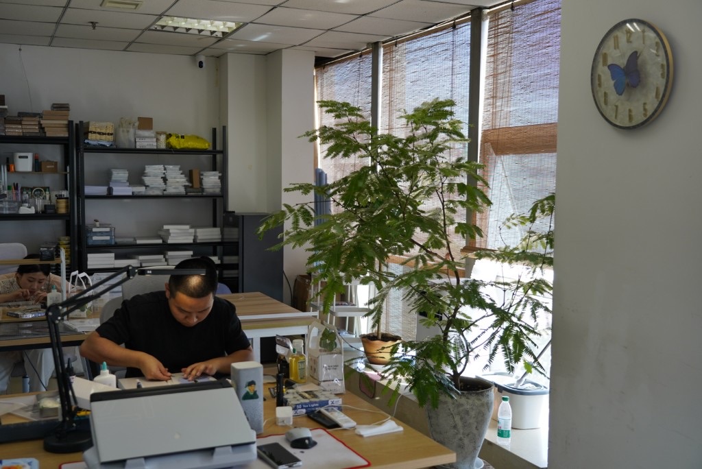 靠窗工位与竹帘采光，工匠在木质桌前作业，旁侧物料架与绿植