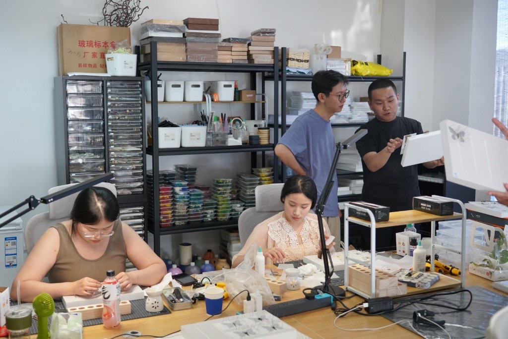 山海造物工坊全景：多人在木质工作台前组装标本匣，货架上码放框架与物料